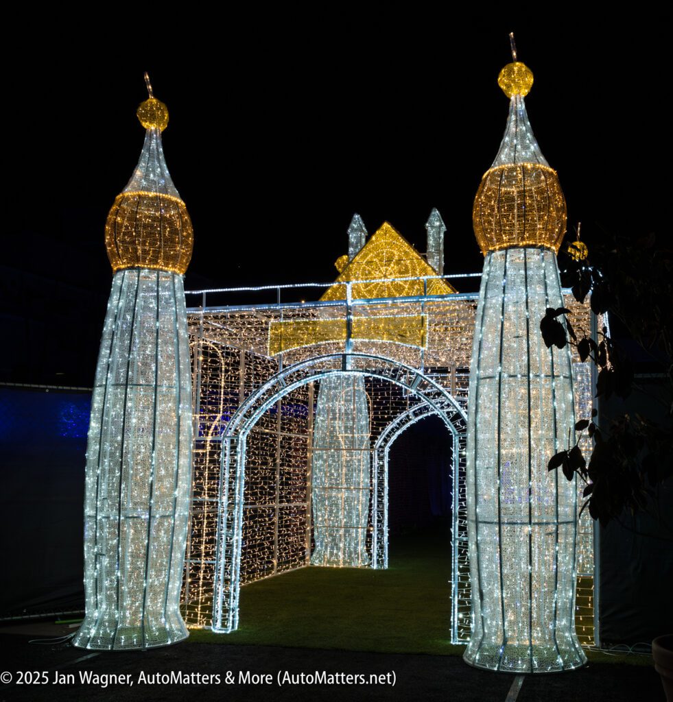 Illuminated structure resembling a castle gate with two tall towers and gold domes, decorated with white and yellow lights, displayed at night.