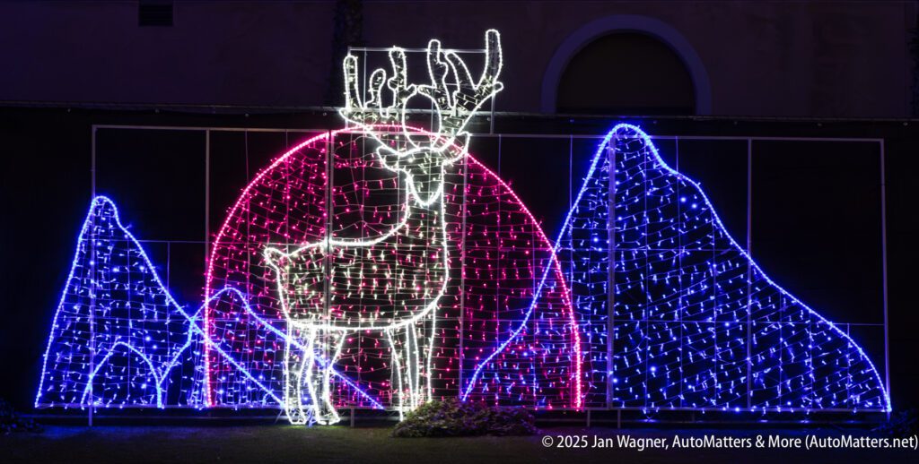 A large light display depicts a reindeer in front of a pink semicircle, with blue mountains made of lights on either side, set against a dark background.
