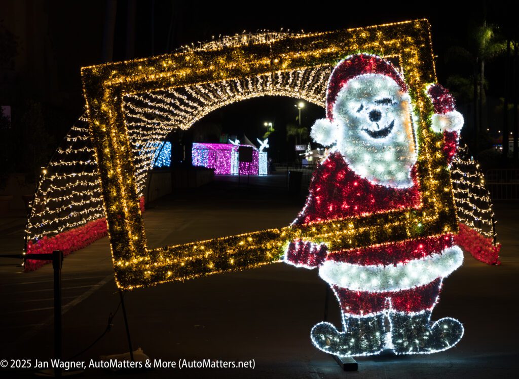 A light display featuring a smiling Santa Claus figure holding a rectangular frame, with more illuminated decorations visible in the background at night.