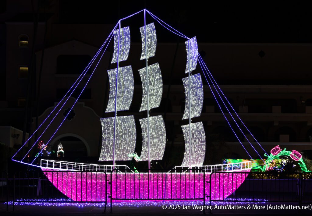 A large ship-shaped holiday light display with white and pink lights stands outdoors at night, with additional illuminated decorations in the background.