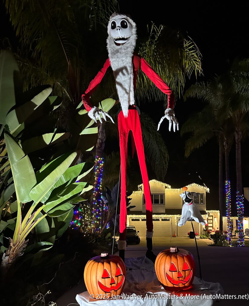 A tall Jack Skellington figure in a red suit stands outdoors at night, surrounded by palm trees, jack-o'-lanterns, and holiday lights, with a small ghost dog nearby.