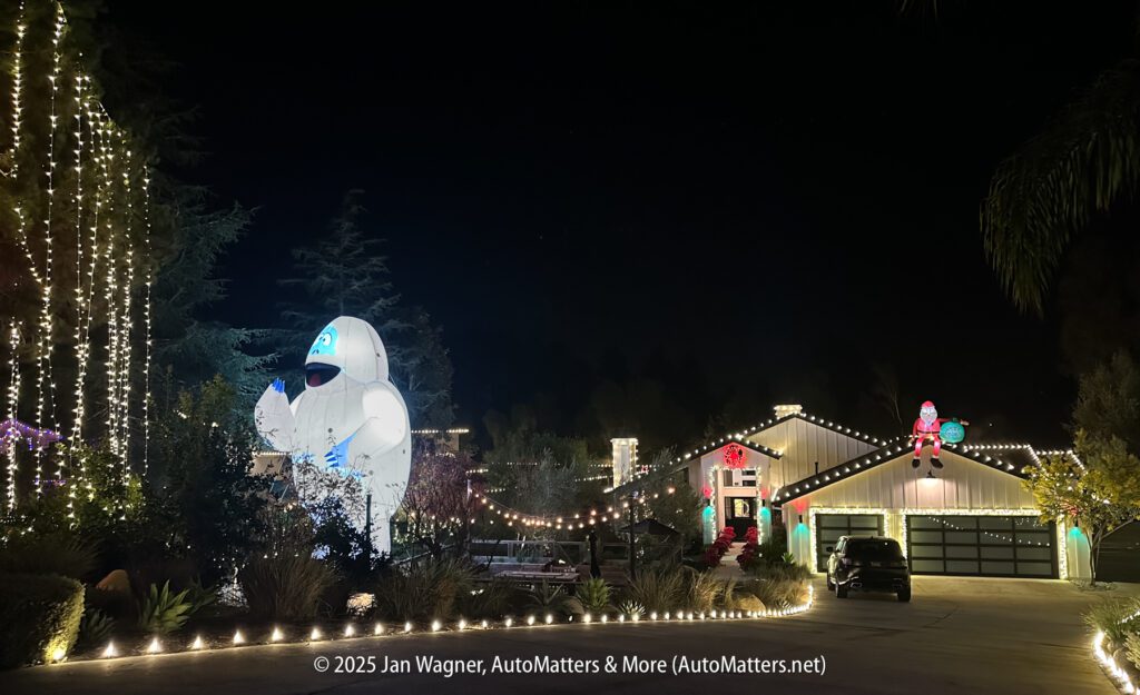 A house and yard decorated with holiday lights and large inflatable characters at night. Trees and shrubs are also lit, and a driveway leads to the garage.