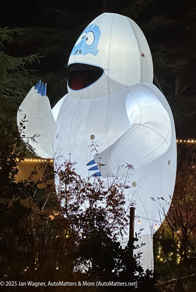 A large inflatable snow monster with blue features and an open mouth is lit up at night, standing among trees and bushes.