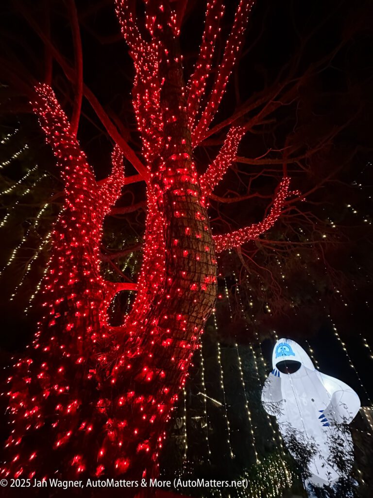 A tree wrapped in red lights stands next to an illuminated white inflatable figure with blue facial features at night, with string lights in the background.