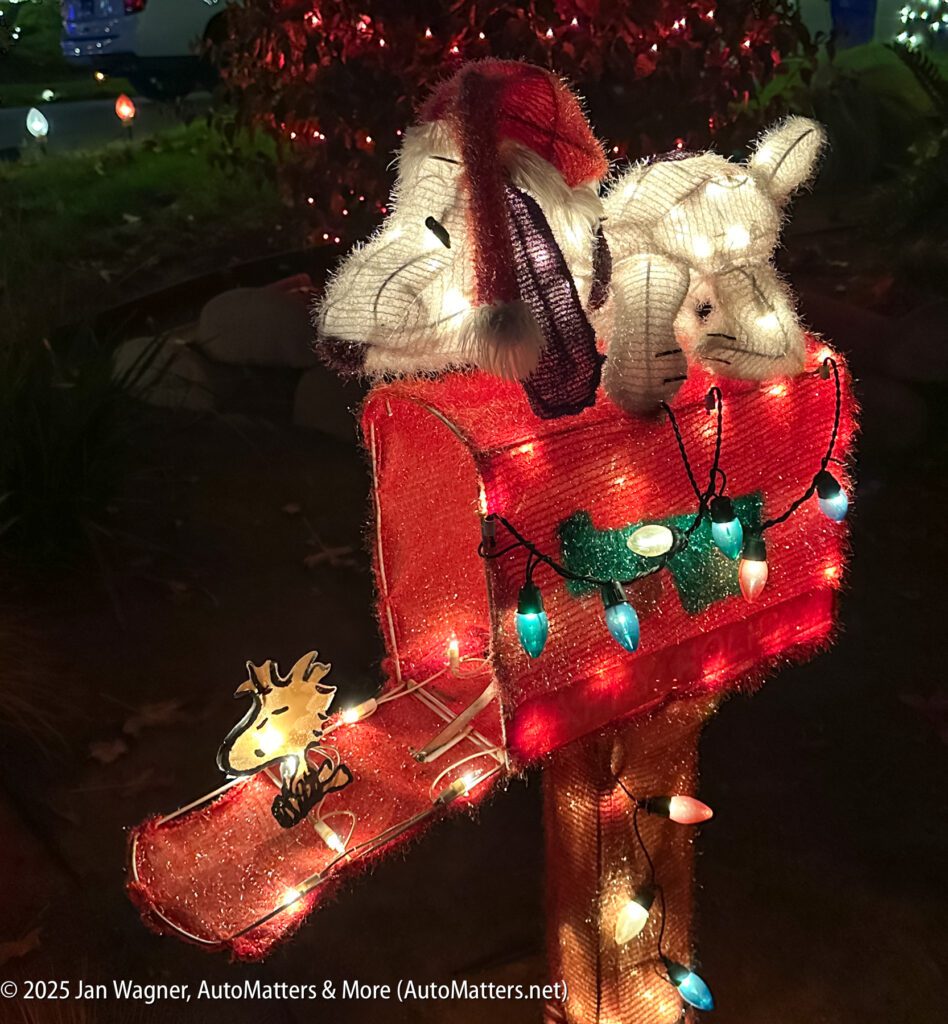 A red mailbox decorated with Christmas lights features plush figures of Snoopy and Woodstock in holiday attire.