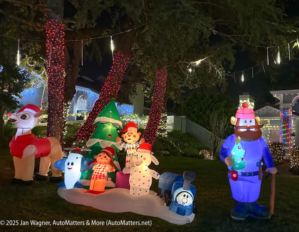 Inflatable Christmas decorations, including Santa, snowmen, a train, and a bear, are displayed outdoors at night with trees and a house lit by string lights.
