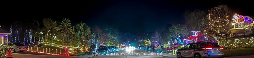 A suburban street at night decorated with colorful holiday lights on houses, trees, and lawns; several cars are parked along the road.
