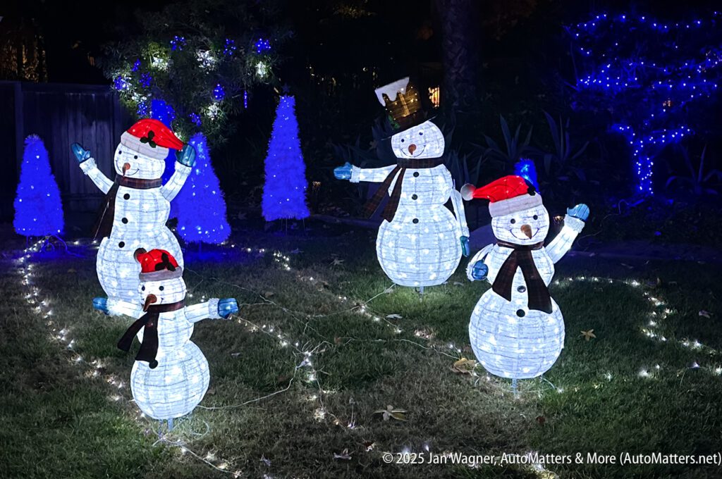 Four illuminated snowman decorations with red hats and scarves are displayed in a yard at night, surrounded by blue-lit trees and festive lights.