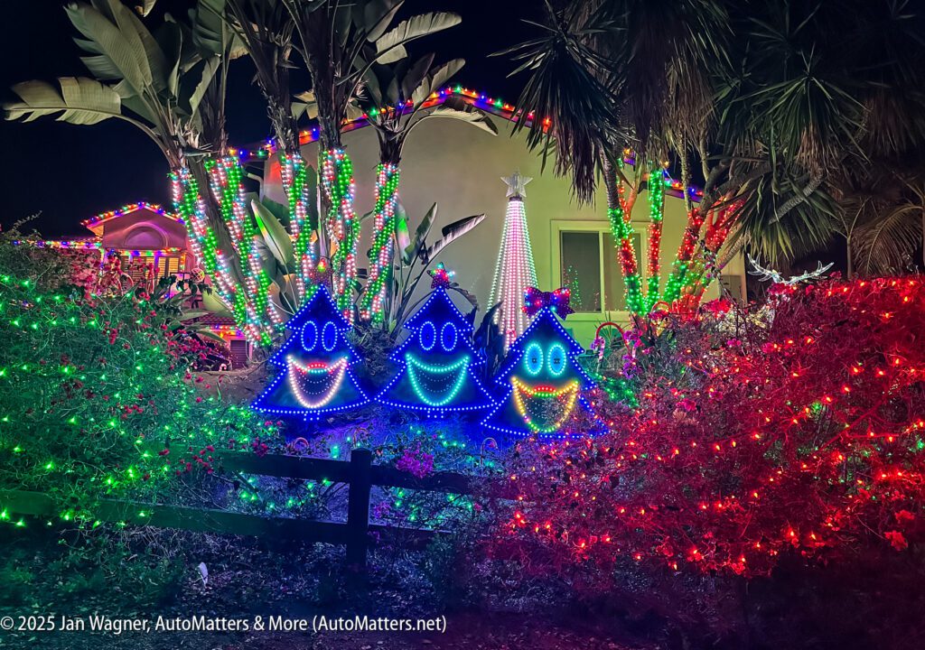 A house and garden decorated with colorful Christmas lights, featuring three large, smiling Christmas tree figures lit in blue, green, and red.