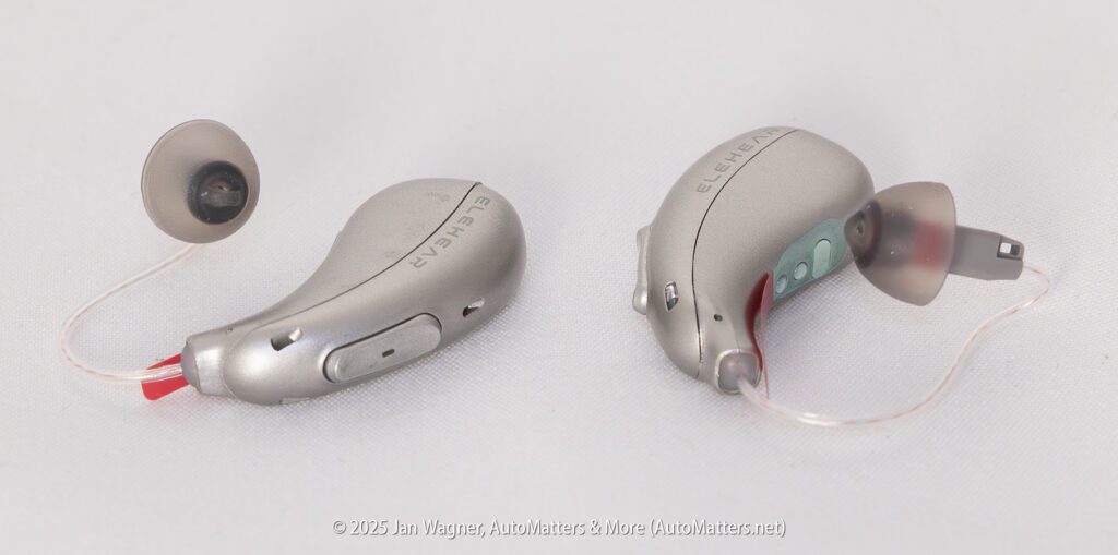 Two silver Elac Hearing hearing aids with attached ear domes and thin tubing are shown on a white surface.