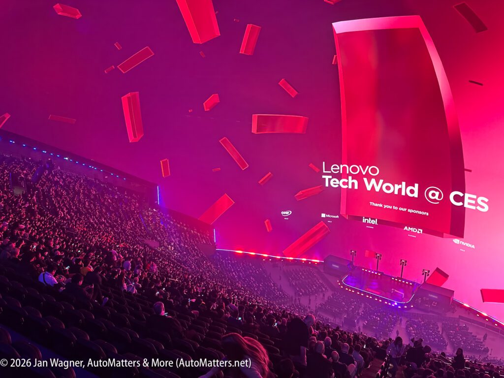 Large audience seated in a futuristic, red-lit auditorium at Lenovo Tech World @ CES; stage displays event sponsors and technology branding.