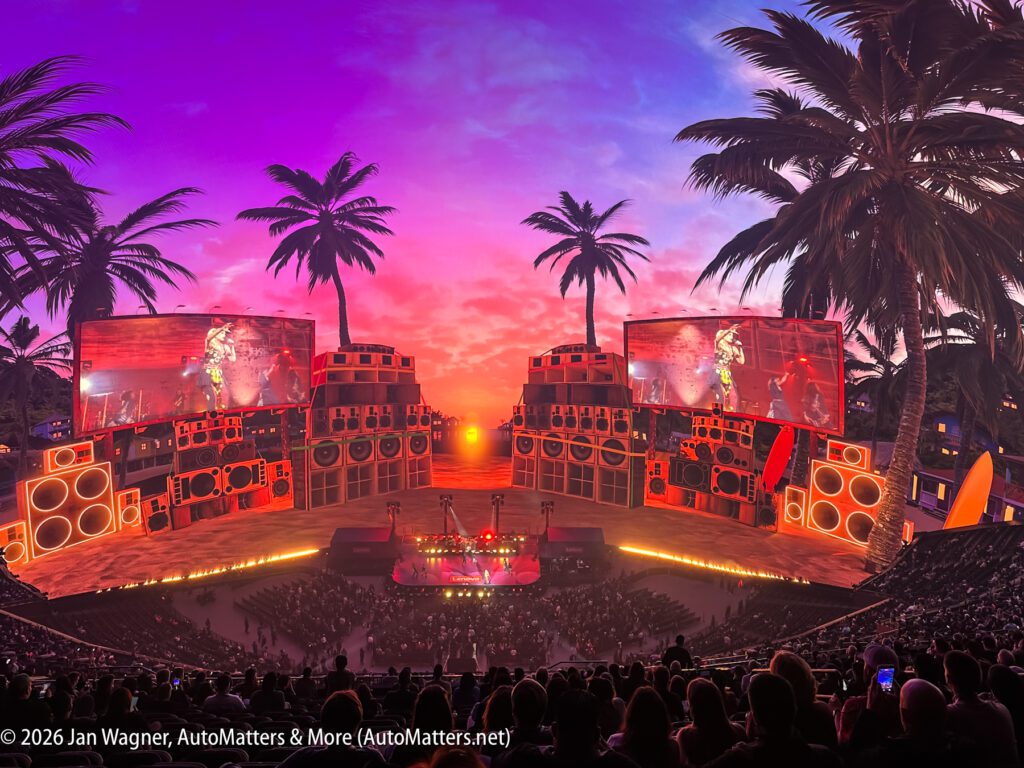 Wide-angle view of a concert stage with tropical-themed decor, large video screens, palm trees, vivid sunset lighting, and an audience seated in front.