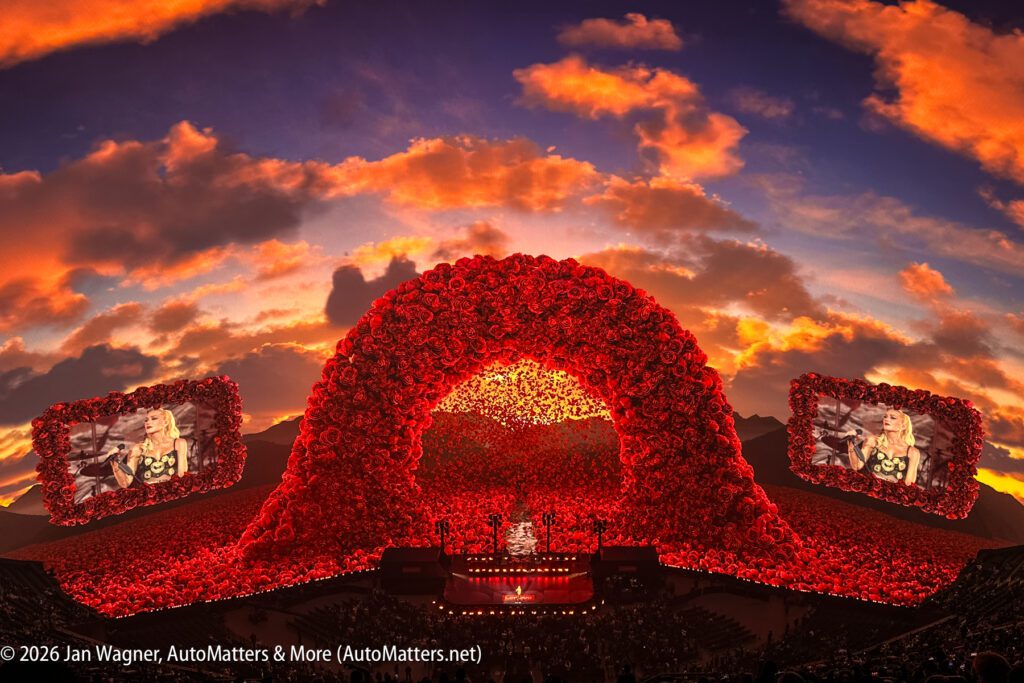 A large outdoor concert stage decorated with red roses features a central arch and two large screens, set against a dramatic sunset sky with clouds.