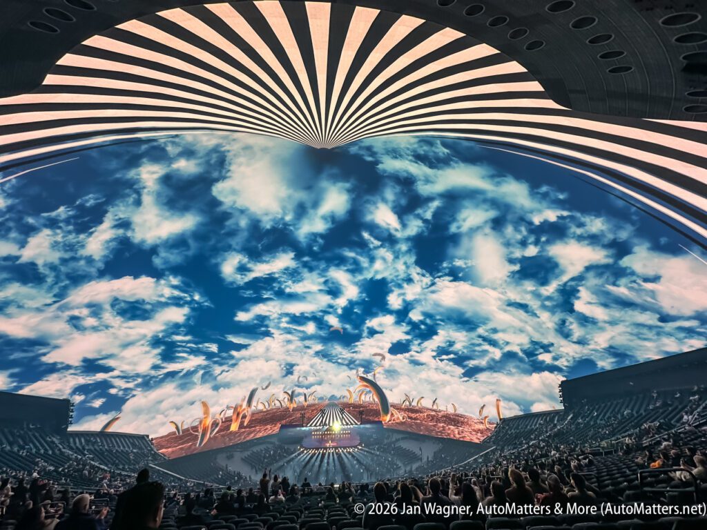 A large indoor arena with a curved ceiling displays a vivid sky scene with clouds; an audience sits below, facing a stage with abstract decorations.