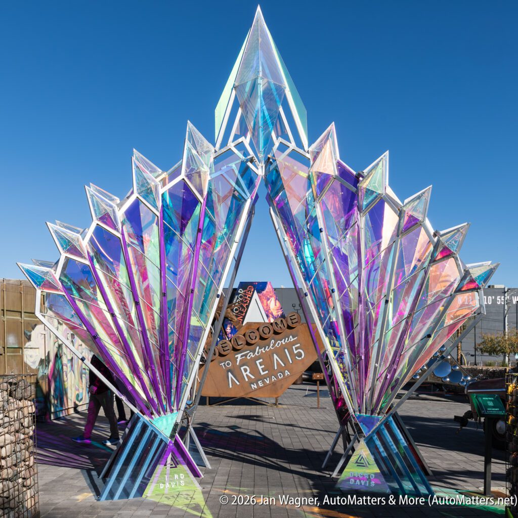 A geometric, iridescent art installation stands outdoors with a “Welcome to Fabulous Area 15 Nevada” sign displayed in the center. Several people are visible around the structure.