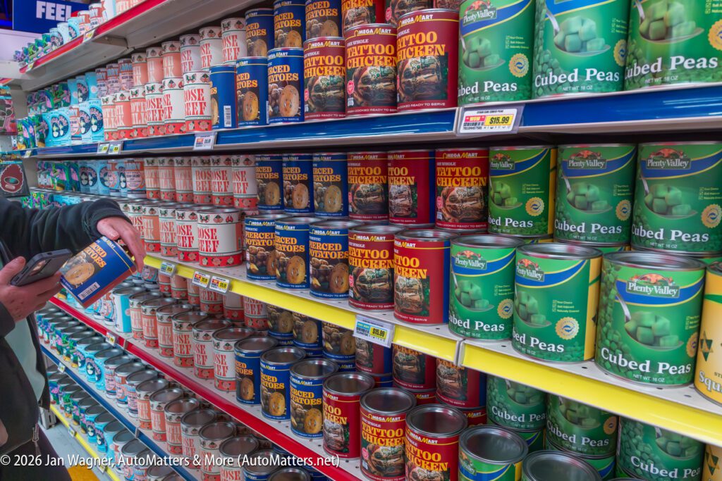 A person holds a can while shopping in a grocery store aisle stocked with various canned goods, including chicken and peas.