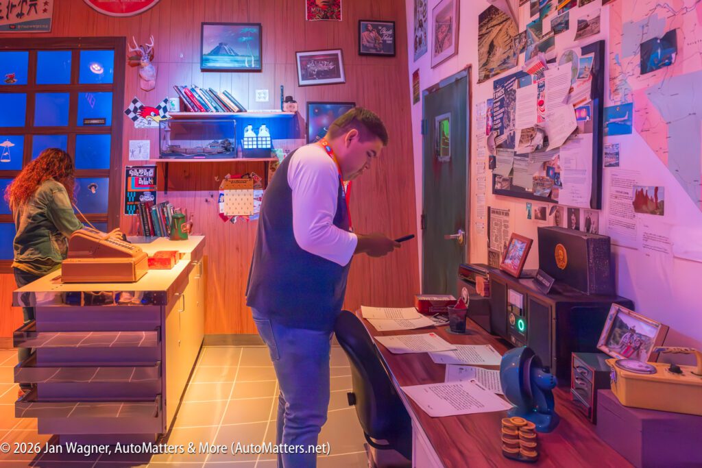 Two people work in a cluttered office full of papers, maps, and vintage items; one stands using a phone, the other sits at a desk with a typewriter.