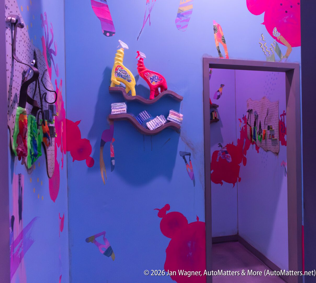 A brightly painted room with blue walls, cleaning supplies, towels on a shelf, and hooks with gloves, featuring colorful abstract wall art.
