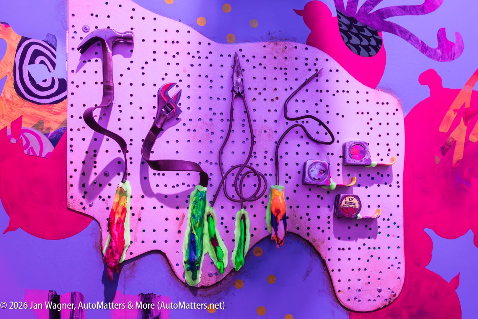A purple pegboard shaped like a paint palette holds tools including a wrench, pliers, saw, tape measures, and paintbrushes against a colorful wall with abstract shapes.