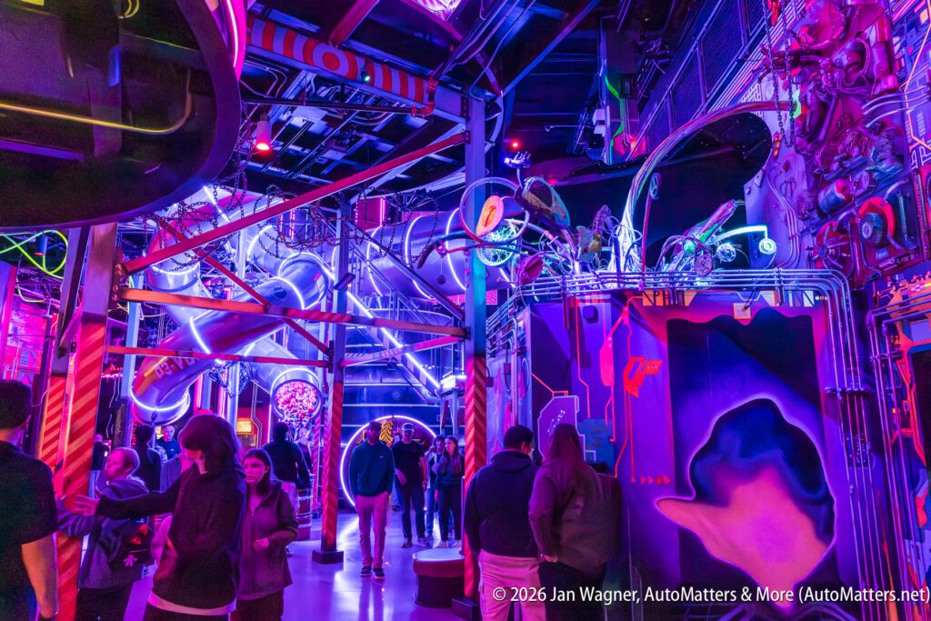 People walk through a vibrant, neon-lit indoor space filled with futuristic decorations, pipes, and colorful lights.