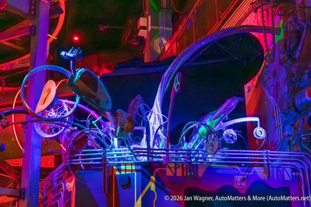 Colorful, illuminated sculptures made from various recycled mechanical parts and bicycles are displayed in an indoor setting with vibrant lighting and industrial elements.