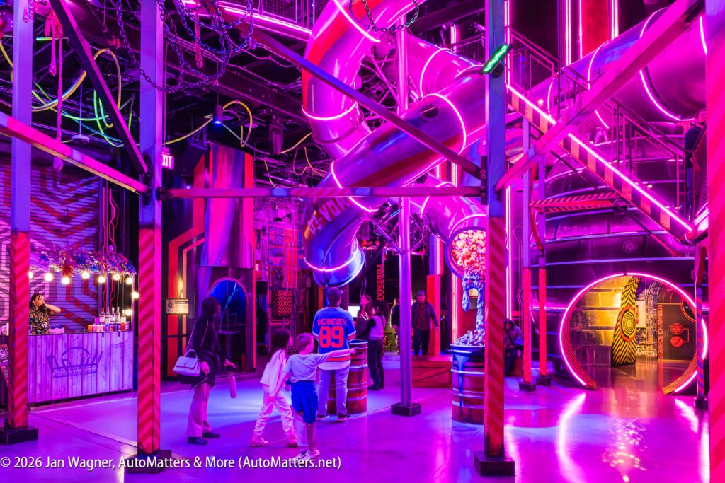 People walk through a vibrant indoor space with neon purple and pink lights, metal structures, and a large tube slide. A concession stand is visible on the left side.