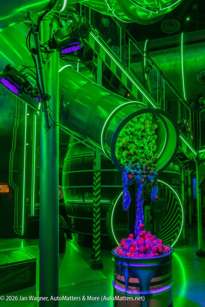A large metallic tube pours a mix of green and red artificial objects under glowing green lights in a futuristic, industrial-themed room.