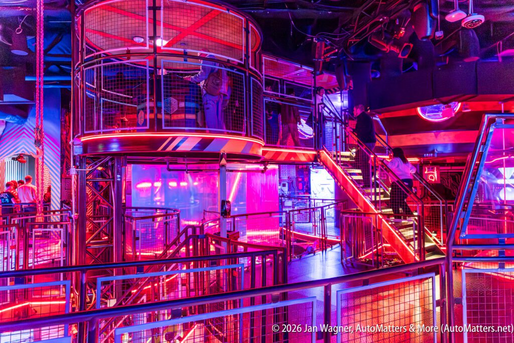 A modern, multi-level indoor venue with vibrant purple and pink neon lighting, metal railings, staircases, and people standing on different levels.