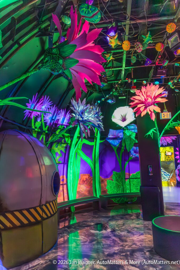 A colorful, neon-lit room features oversized artificial flowers, curved benches, a small metallic structure, and an exit sign.