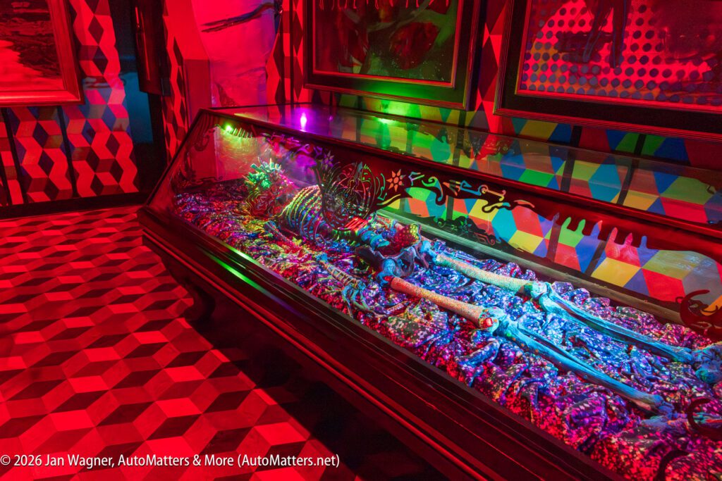 A human skeleton displayed in a glass case with vibrant, multicolored lighting and patterned walls and floor, creating a surreal atmosphere.
