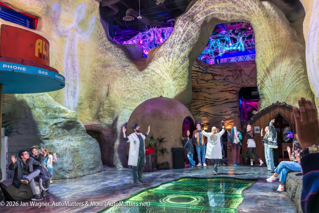 People in lab coats perform for an audience inside a colorful, cave-themed, interactive exhibit with projected lights and sci-fi decor.