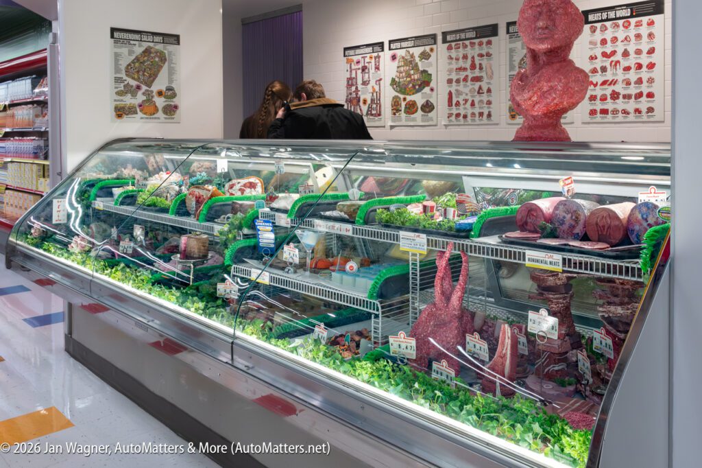 A refrigerated display case filled with various cuts of raw meat in a supermarket; two people are selecting items, and meat charts are visible on the wall behind the counter.