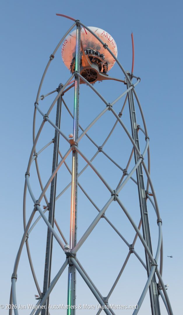 A spherical amusement ride capsule ascends a tall, spiral metal tower structure against a clear blue sky.