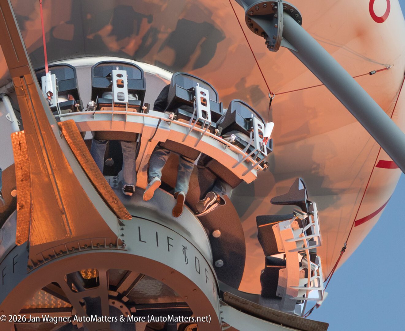Passengers seated in a circular amusement park ride as it ascends, with legs dangling and safety restraints secured.