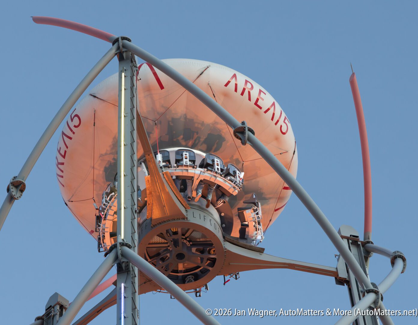 Close-up view of a futuristic amusement ride labeled “ARELIS,” showing its central mechanism and support structure against a clear sky.