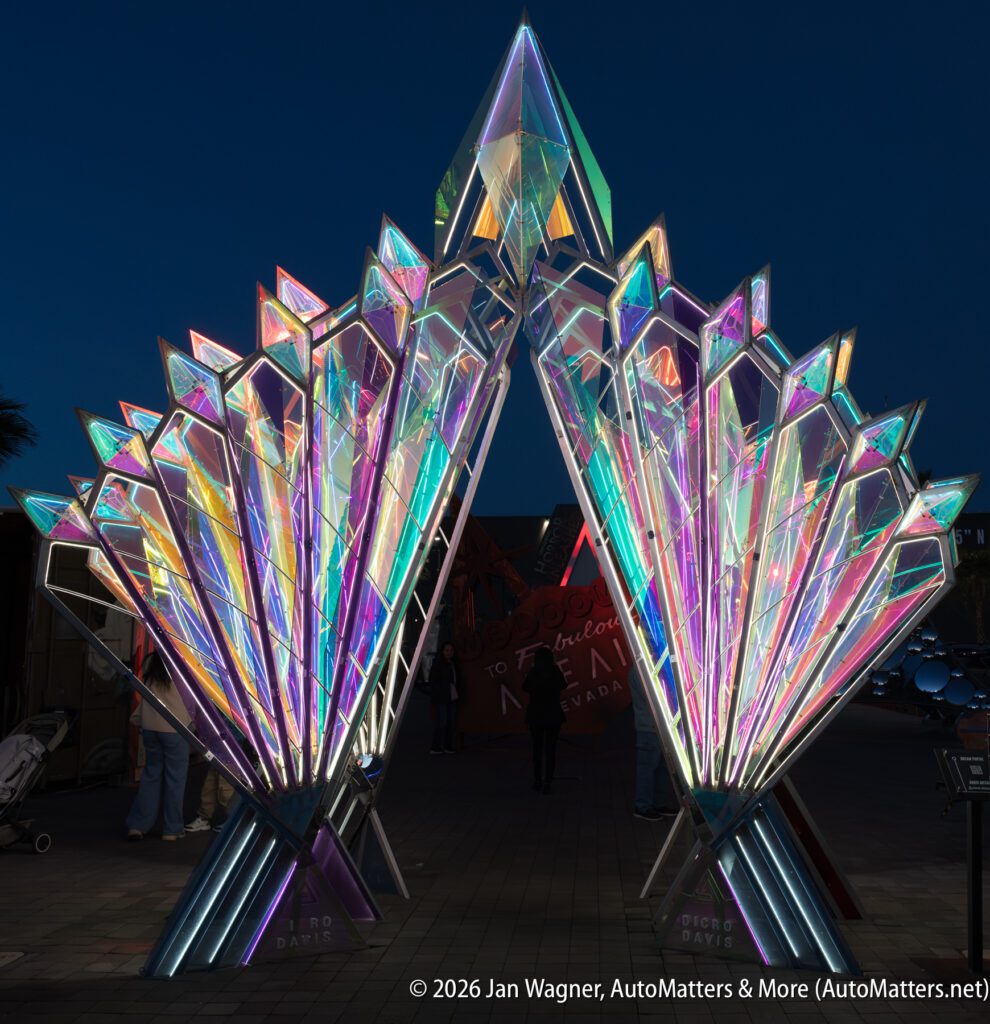 Large illuminated geometric sculpture with multicolored lights, forming an archway at night. People walk around and through the structure.