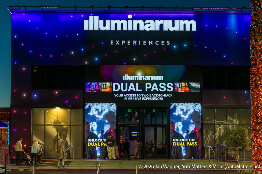 The Illuminarium Experiences building exterior at night, with brightly lit signage and digital screens advertising a dual pass for immersive experiences.