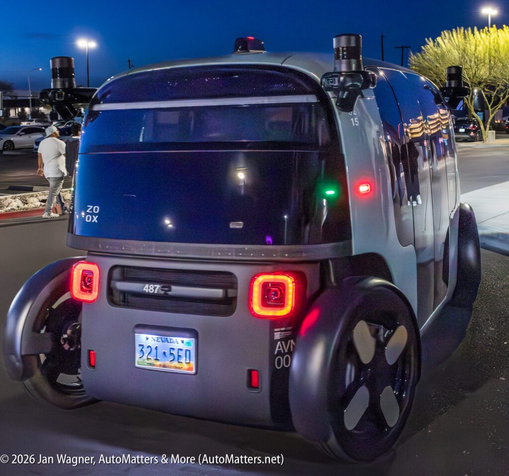 A self-driving, box-shaped autonomous vehicle with Nevada plates is parked on a street at dusk; sensors and cameras are visible on the roof and corners.