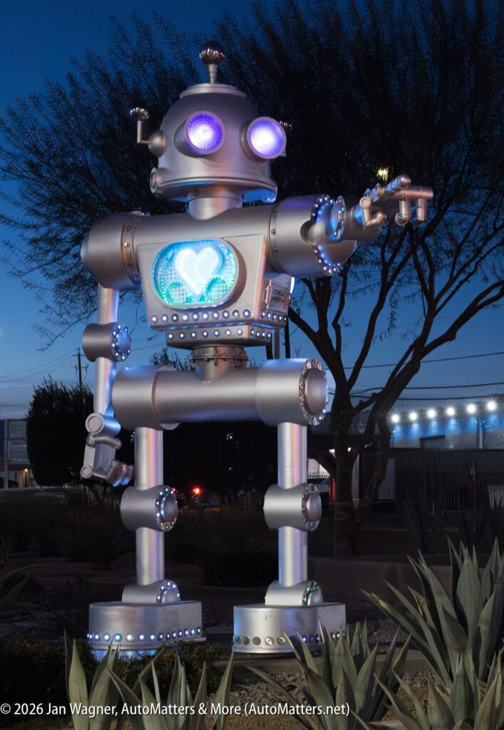 Large silver robot sculpture with blue and white lights stands outdoors at dusk, featuring a heart shape on its chest and one arm extended forward.