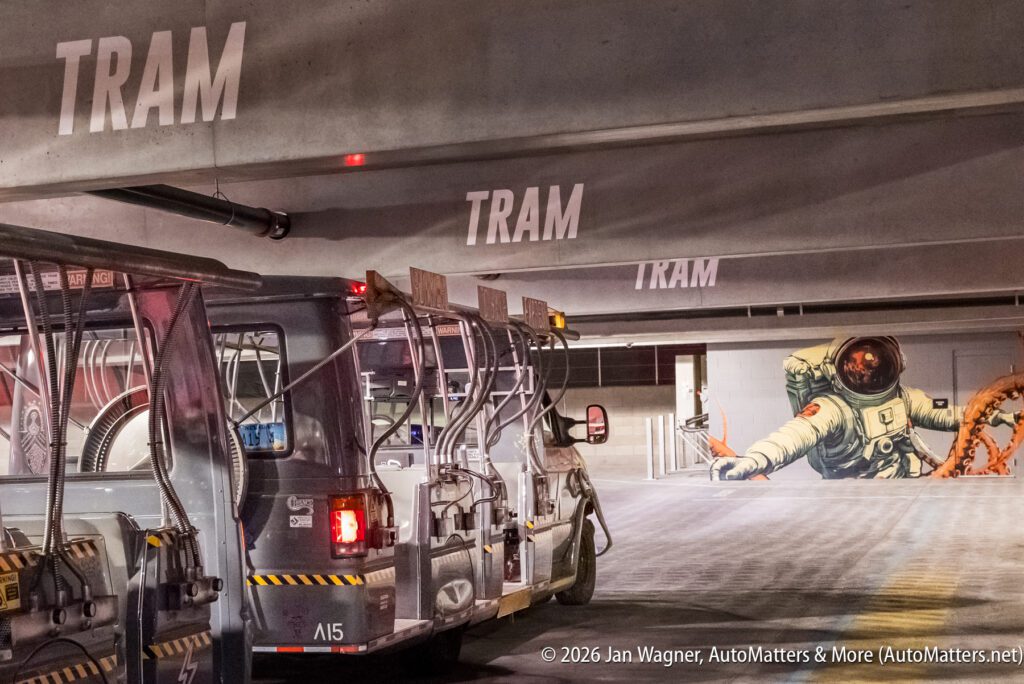 A tram parked in a multi-level garage near a mural of an astronaut with an orange tentacle reaching toward them. "TRAM" is painted on the garage walls.