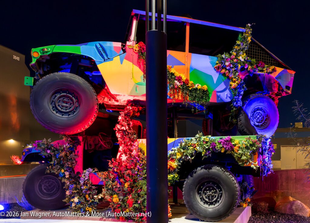 Two off-road vehicles, brightly painted and covered in artificial flowers, are stacked on top of each other and lit with colorful lights at night.