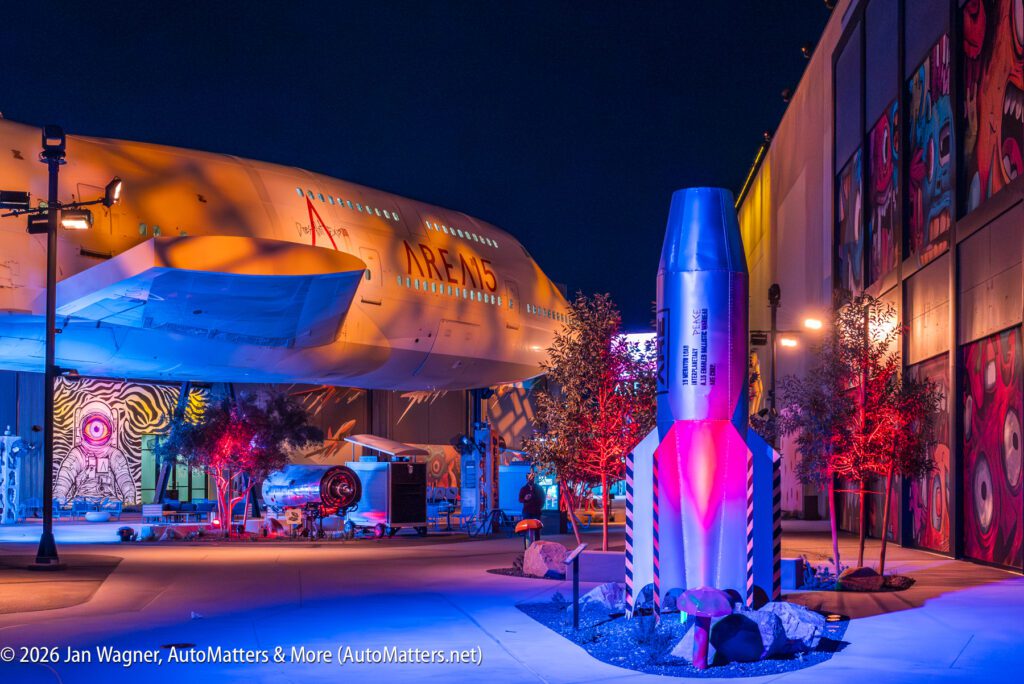 Outdoor nighttime scene showing a large airplane labeled "AREMS" next to a small rocket and colorful mural-covered walls illuminated by vibrant lights.