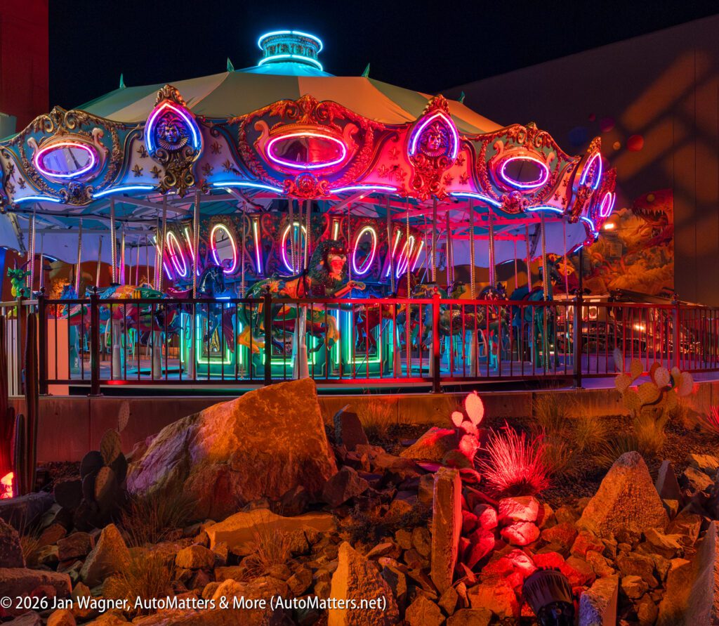 A brightly lit carousel with neon "Welcome" sign is seen at night, surrounded by desert rocks and cacti in the foreground.