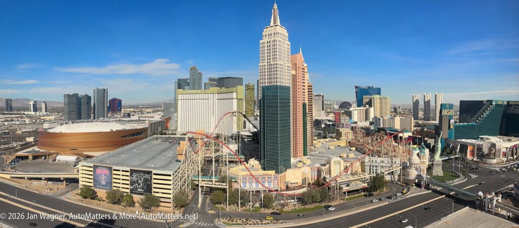 Panoramic view of the Las Vegas Strip featuring hotels, casinos, a roller coaster, city buildings, and distant mountains under a clear blue sky.