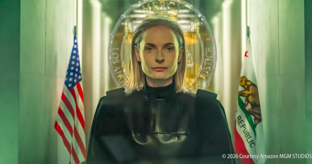 A person with shoulder-length gray hair stands in front of U.S. and California flags, wearing a black outfit, with an official seal in the background.