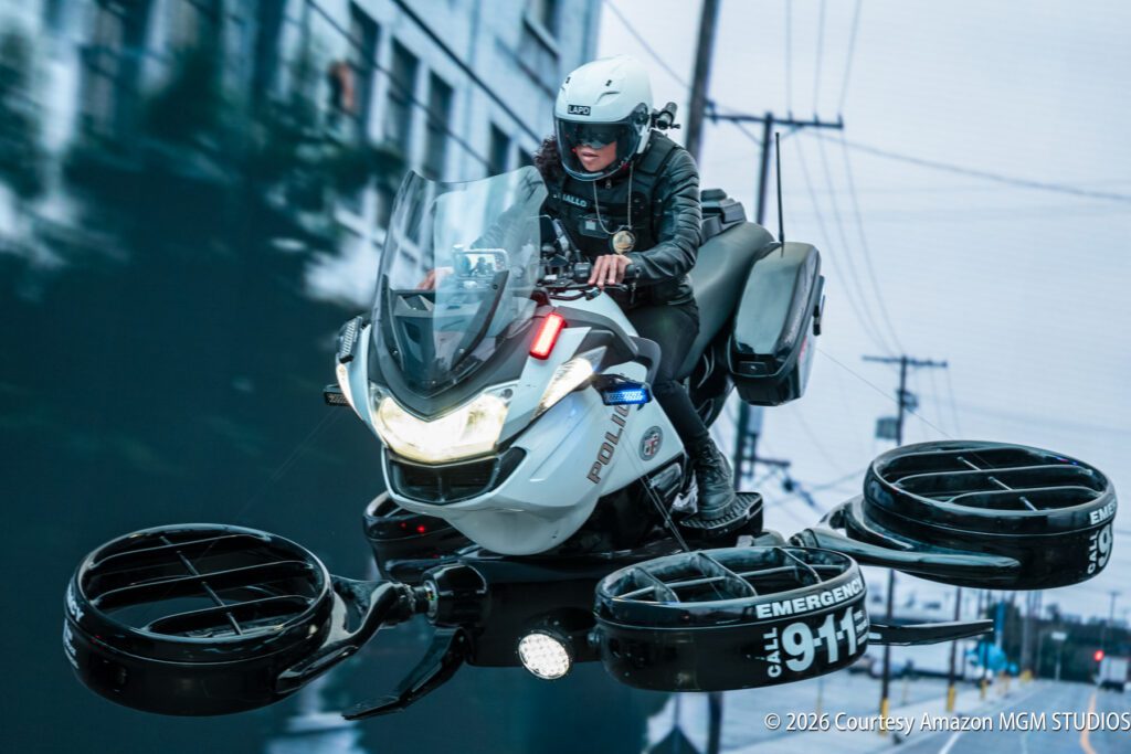 A police officer rides a futuristic hovering vehicle above a city street, equipped with emergency lights and labeled with "911" and "POLICE.