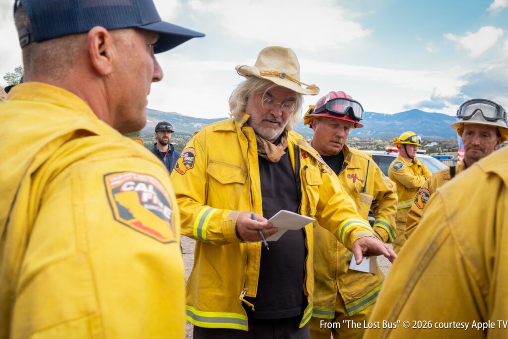 A man in a yellow firefighter jacket and straw hat speaks to a group of firefighters outdoors, holding papers; mountains are visible in the background.