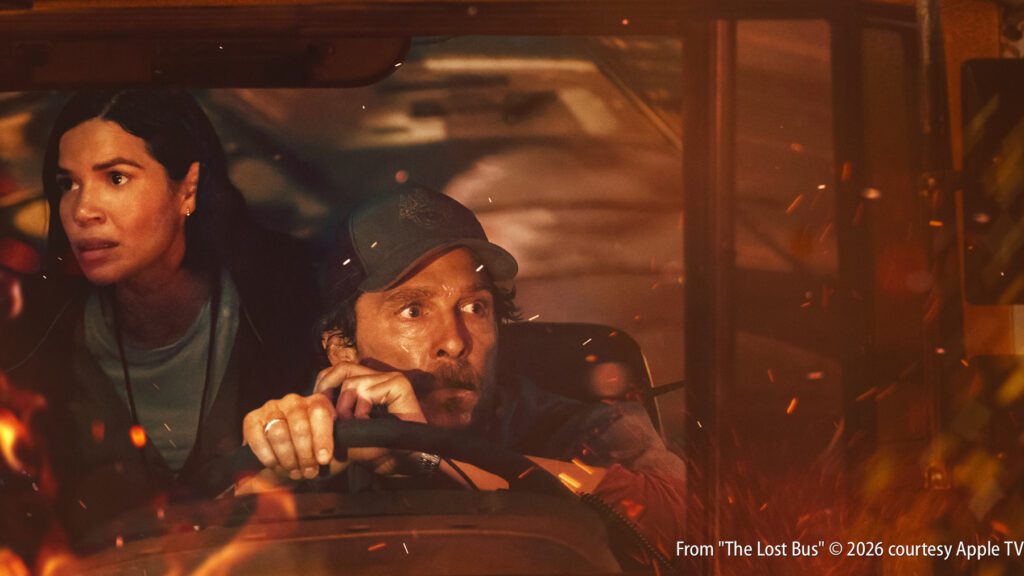 A man drives a vehicle through a scene with fire and flying embers, while a concerned woman sits beside him.