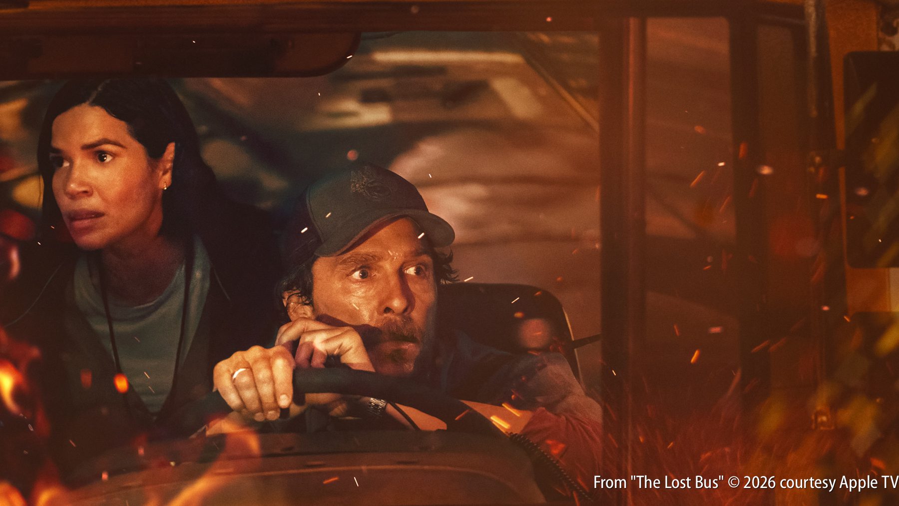 A man drives a vehicle through a scene with fire and flying embers, while a concerned woman sits beside him.