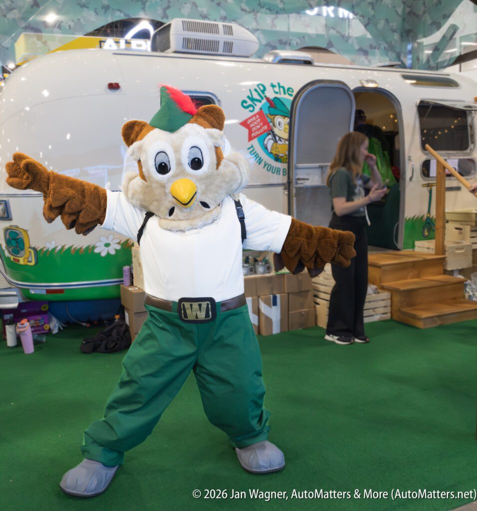 A person in an owl mascot costume poses with outstretched arms in front of a trailer, while another person stands nearby.